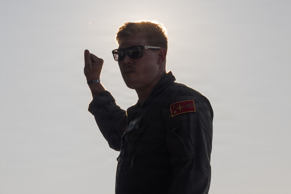 A young male in a dark military-style flight suit is photographed against a bright backlit sky, creating a partial silhouette effect. He is wearing sunglasses and has a patch on his left shoulder that reads 'PC' on a red and gold background. He is raising his right fist/hand in what appears to be a gesture or signal.