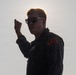 A young male in a dark military-style flight suit is photographed against a bright backlit sky, creating a partial silhouette effect. He is wearing sunglasses and has a patch on his left shoulder that reads 'PC' on a red and gold background. He is raising his right fist/hand in what appears to be a gesture or signal.