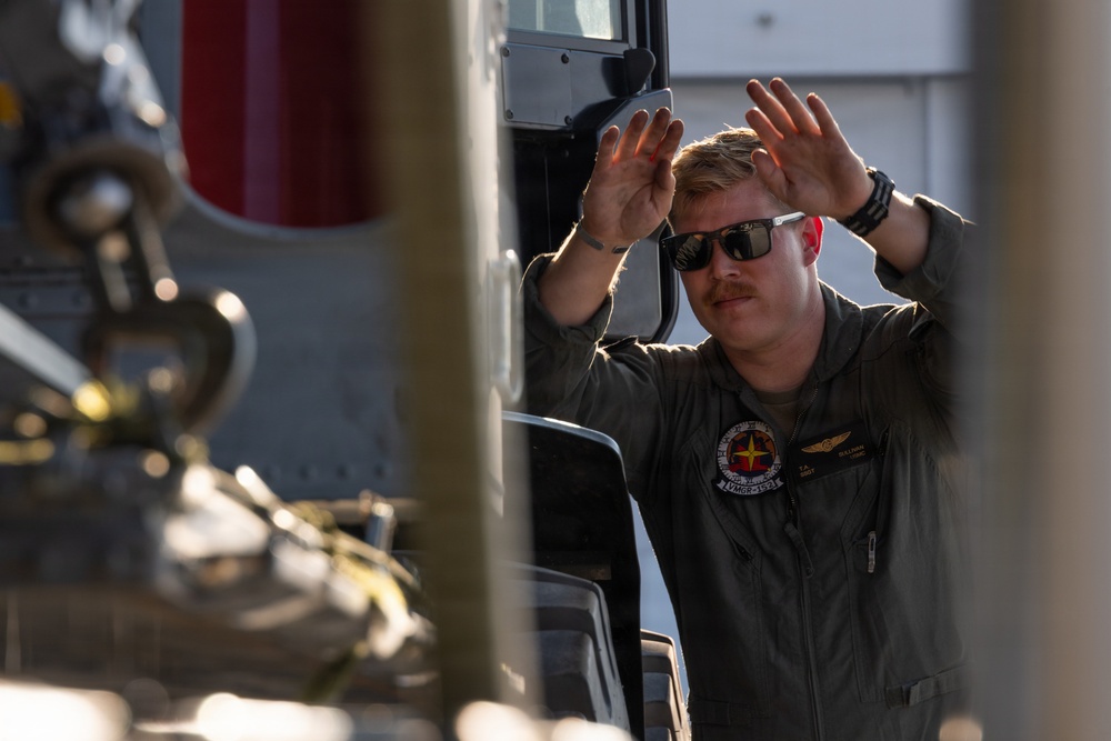 A military aviator or crew member in an olive/sage green flight suit is standing beside what appears to be a military helicopter, raising both hands in a signaling or marshaling gesture. The individual is wearing sunglasses, has a mustache, and displays a squadron patch on their chest. The image is taken from a partially obscured angle with helicopter components visible in the foreground.
