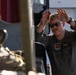 A military aviator or crew member in an olive/sage green flight suit is standing beside what appears to be a military helicopter, raising both hands in a signaling or marshaling gesture. The individual is wearing sunglasses, has a mustache, and displays a squadron patch on their chest. The image is taken from a partially obscured angle with helicopter components visible in the foreground.