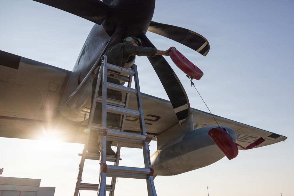 A maintenance technician is standing on an aluminum ladder performing work on a multi-blade propeller of a military turboprop aircraft. Red protective covers (intake/exhaust plugs) are visible attached to the engine nacelle and wing-mounted pod. The image is taken from a low angle at what appears to be dusk or dawn, with strong backlighting from the sun.