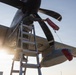 A maintenance technician is standing on an aluminum ladder performing work on a multi-blade propeller of a military turboprop aircraft. Red protective covers (intake/exhaust plugs) are visible attached to the engine nacelle and wing-mounted pod. The image is taken from a low angle at what appears to be dusk or dawn, with strong backlighting from the sun.