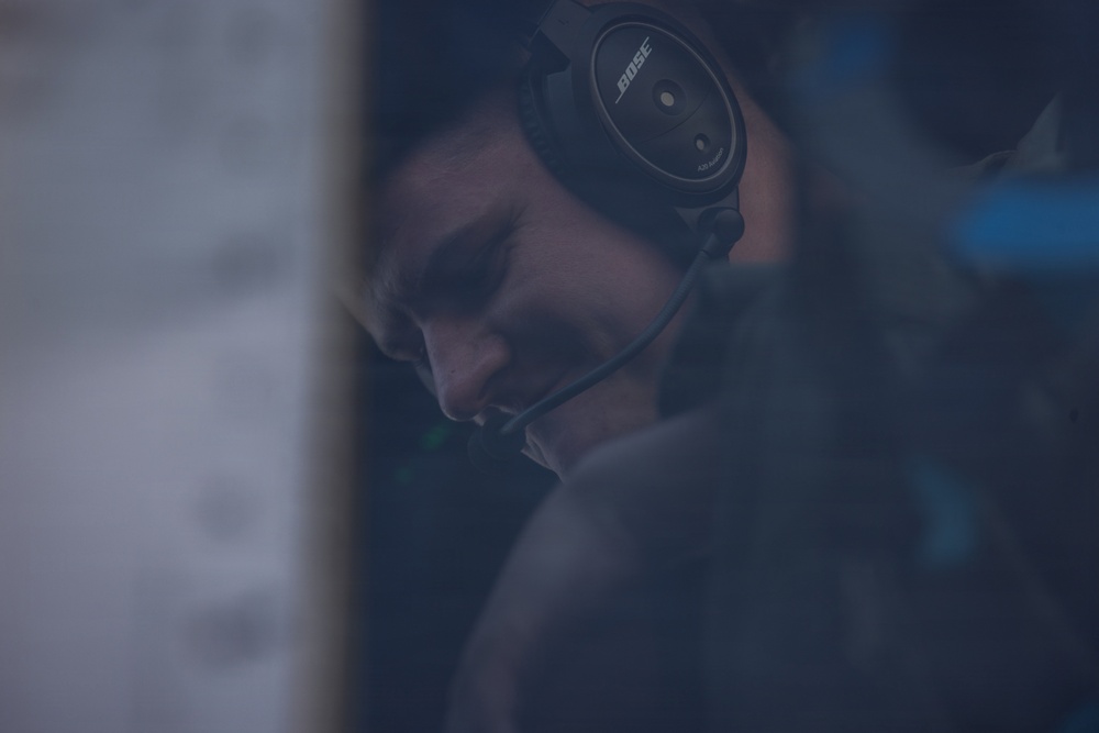 A person wearing a Bose aviation/professional headset with a boom microphone is visible in a dimly lit, moody environment. The subject appears to be focused downward, possibly monitoring or communicating in a professional capacity. The image has a dark, blue-toned aesthetic with shallow depth of field.