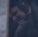 A person wearing a Bose aviation/professional headset with a boom microphone is visible in a dimly lit, moody environment. The subject appears to be focused downward, possibly monitoring or communicating in a professional capacity. The image has a dark, blue-toned aesthetic with shallow depth of field.