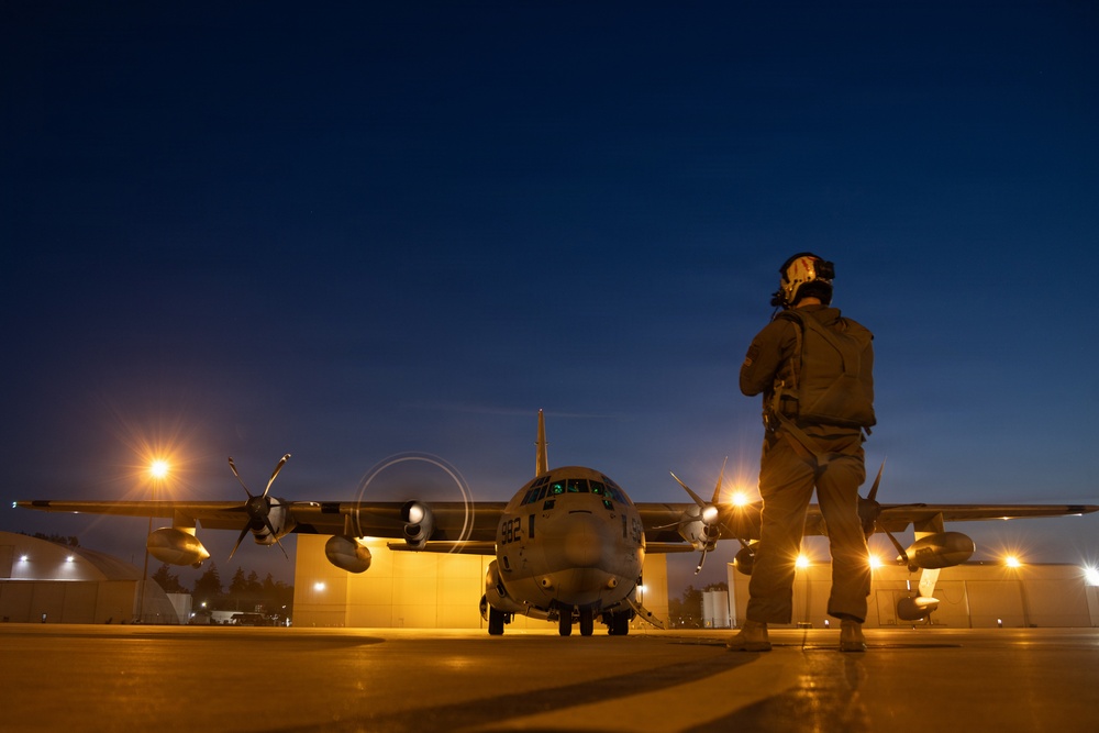 A military flight crew member stands on a tarmac at dusk or dawn observing a four-engine turboprop military transport aircraft, identifiable as a C-130 Hercules variant, with tail number partially visible as '382'. The scene is illuminated by warm ground lighting against a deep blue twilight sky, creating a dramatic silhouette effect. The aircraft appears to be undergoing a pre-flight or post-flight inspection on what appears to be a military airfield.