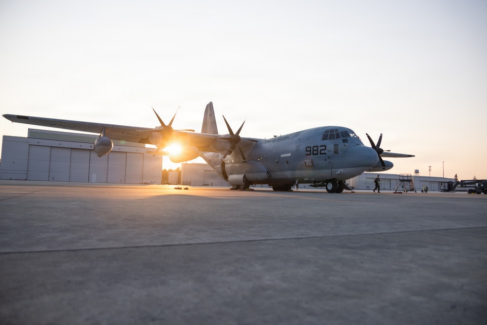 A military turboprop transport aircraft marked '982' is parked on a concrete flight line apron at what appears to be a military air station, photographed during golden hour with the sun creating a lens flare behind the aircraft. The aircraft is a C-130 Hercules variant in gray livery, consistent with U.S. Navy or Marine Corps markings. Ground crew personnel and support equipment are visible in the background near a large hangar.