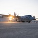 A military turboprop transport aircraft marked '982' is parked on a concrete flight line apron at what appears to be a military air station, photographed during golden hour with the sun creating a lens flare behind the aircraft. The aircraft is a C-130 Hercules variant in gray livery, consistent with U.S. Navy or Marine Corps markings. Ground crew personnel and support equipment are visible in the background near a large hangar.