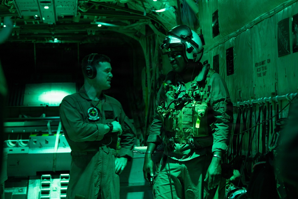 Two military personnel are inside the cargo hold of a military aircraft, illuminated by green lighting typical of night operations. One individual wears a flight suit with headset and unit patch, while the other is fully equipped with tactical gear, helmet, night vision mount, and body armor. They appear to be in conversation, likely during a pre-jump or mission briefing.