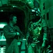 Two military personnel are inside the cargo hold of a military aircraft, illuminated by green lighting typical of night operations. One individual wears a flight suit with headset and unit patch, while the other is fully equipped with tactical gear, helmet, night vision mount, and body armor. They appear to be in conversation, likely during a pre-jump or mission briefing.