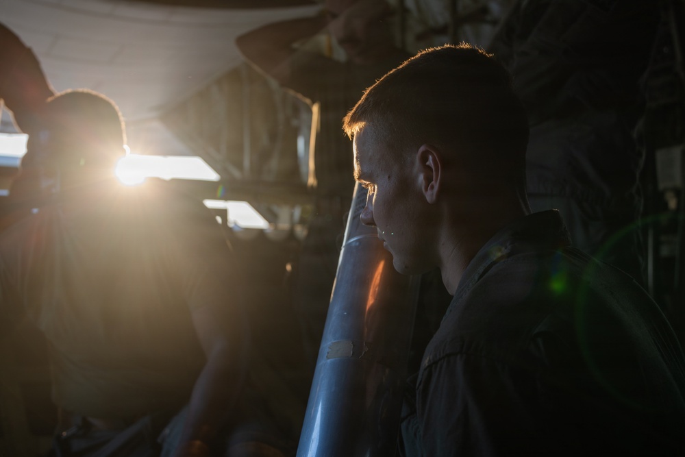 A young male individual is photographed in profile in a dimly lit industrial or mechanical environment, with a strong backlight source creating dramatic lens flare on the left side of the frame. A cylindrical metallic object is visible in the foreground near the subject. The lighting conditions create a high-contrast, moody aesthetic with deep shadows.