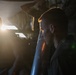 A young male individual is photographed in profile in a dimly lit industrial or mechanical environment, with a strong backlight source creating dramatic lens flare on the left side of the frame. A cylindrical metallic object is visible in the foreground near the subject. The lighting conditions create a high-contrast, moody aesthetic with deep shadows.