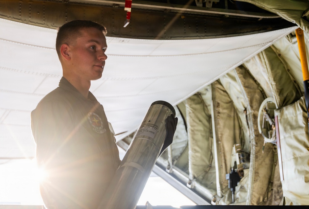 A young military personnel member in a flight suit is holding a cylindrical metallic canister inside what appears to be a military aircraft cargo bay. The individual is illuminated by strong backlighting, creating a dramatic silhouette effect. The interior of the aircraft shows olive-drab fabric lining, structural ribs, and various equipment attachments.