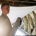 A young military personnel member in a flight suit is holding a cylindrical metallic canister inside what appears to be a military aircraft cargo bay. The individual is illuminated by strong backlighting, creating a dramatic silhouette effect. The interior of the aircraft shows olive-drab fabric lining, structural ribs, and various equipment attachments.