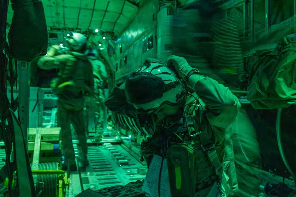Military personnel are photographed inside what appears to be a large military transport aircraft, illuminated by green lighting consistent with night operations. The individuals are wearing full tactical gear including helmets, parachute harnesses, and combat equipment, suggesting preparation for a airborne insertion or jump operation. The motion blur in the image indicates movement and activity within the aircraft cabin.