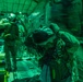 Military personnel are photographed inside what appears to be a large military transport aircraft, illuminated by green lighting consistent with night operations. The individuals are wearing full tactical gear including helmets, parachute harnesses, and combat equipment, suggesting preparation for a airborne insertion or jump operation. The motion blur in the image indicates movement and activity within the aircraft cabin.