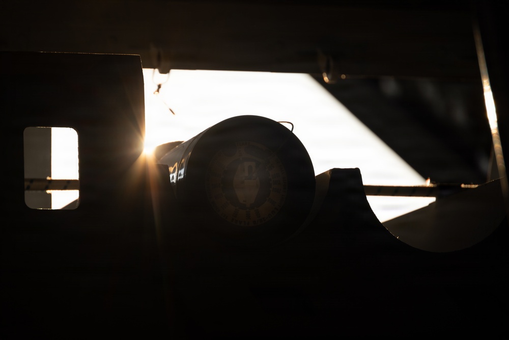 A heavily backlit, silhouetted scene showing what appears to be a military or official vehicle interior/exterior, with a circular emblem or seal visible on a dark surface. Strong sunlight or artificial light sources create dramatic lens flare and rim lighting effects. The extreme contrast between light and shadow renders most details in deep shadow.