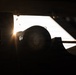 A heavily backlit, silhouetted scene showing what appears to be a military or official vehicle interior/exterior, with a circular emblem or seal visible on a dark surface. Strong sunlight or artificial light sources create dramatic lens flare and rim lighting effects. The extreme contrast between light and shadow renders most details in deep shadow.