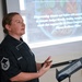 A U.S. Air Force Master Sergeant named Hodges, wearing a black chef's coat, is presenting a cooking and nutrition program to an audience. The presentation slide behind her references empowering Airmen and Sailors with cooking skills supporting the Air Force Comprehensive Airman Fitness Resiliency Pillars. She appears to be speaking at a podium or lectern in an indoor briefing room.