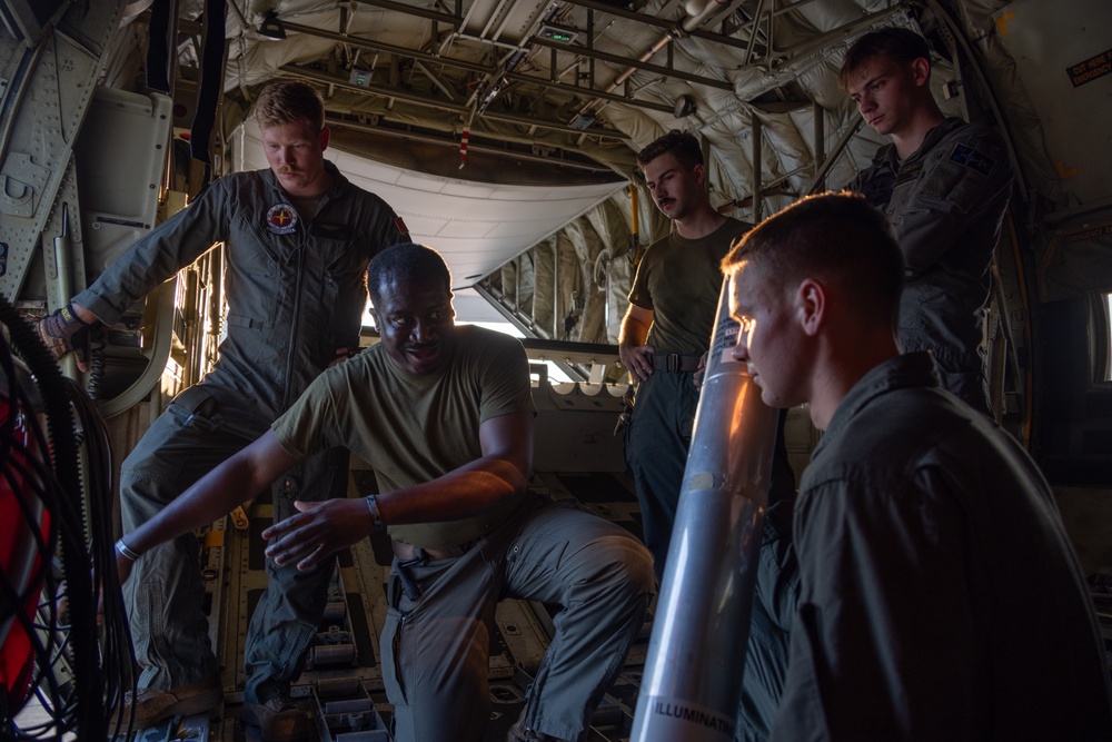 Five military personnel are gathered inside the cargo bay of a large military aircraft, likely a C-130 Hercules. One individual appears to be instructing or briefing the others, with an 'ILLUMINATION' round or flare canister visible in the foreground. The setting suggests a training or mission preparation scenario.