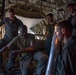 Five military personnel are gathered inside the cargo bay of a large military aircraft, likely a C-130 Hercules. One individual appears to be instructing or briefing the others, with an 'ILLUMINATION' round or flare canister visible in the foreground. The setting suggests a training or mission preparation scenario.