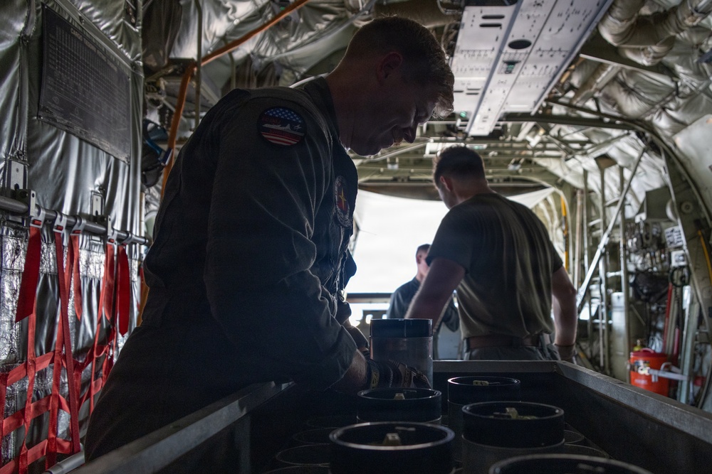 Military personnel are working inside the cargo hold of a large military transport aircraft, likely a C-130 Hercules. A foreground figure wearing an olive flight suit with a U.S. flag patch is handling cylindrical canisters or containers arranged in a row. At least two additional personnel are visible further back in the aircraft's cargo bay.