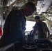Military personnel are working inside the cargo hold of a large military transport aircraft, likely a C-130 Hercules. A foreground figure wearing an olive flight suit with a U.S. flag patch is handling cylindrical canisters or containers arranged in a row. At least two additional personnel are visible further back in the aircraft's cargo bay.
