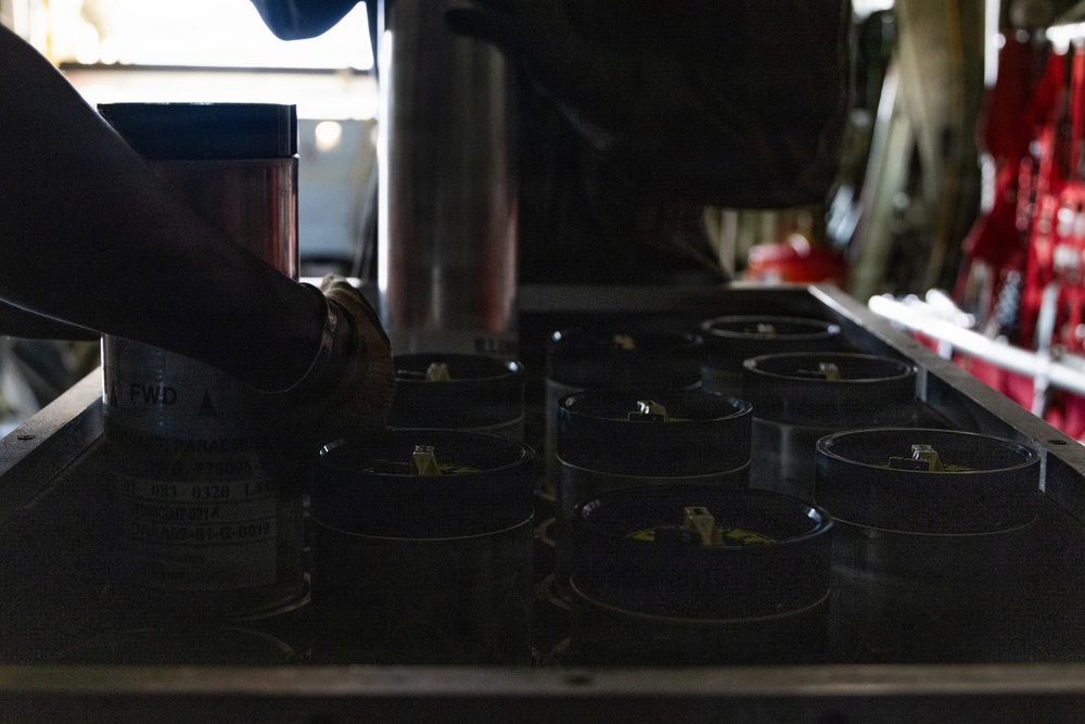 A person is handling cylindrical canisters or dispensers arranged in a grid pattern on a metal tray or rack. The setting appears to be inside a military aircraft or vehicle, suggested by the red equipment visible in the background and the 'FWD' (forward) directional label visible on equipment to the left. The image is dark with limited lighting, making fine details difficult to discern.
