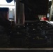 A person is handling cylindrical canisters or dispensers arranged in a grid pattern on a metal tray or rack. The setting appears to be inside a military aircraft or vehicle, suggested by the red equipment visible in the background and the 'FWD' (forward) directional label visible on equipment to the left. The image is dark with limited lighting, making fine details difficult to discern.