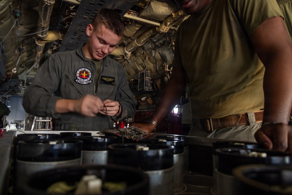 Two military personnel are working inside what appears to be a military aircraft, likely performing maintenance or equipment checks. The individual on the left is wearing a flight suit with unit patches and aviator wings, while the person on the right is in a olive/green t-shirt. They are focused on handling components arranged on a surface in front of them.