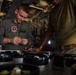 Two military personnel are working inside what appears to be a military aircraft, likely performing maintenance or equipment checks. The individual on the left is wearing a flight suit with unit patches and aviator wings, while the person on the right is in a olive/green t-shirt. They are focused on handling components arranged on a surface in front of them.