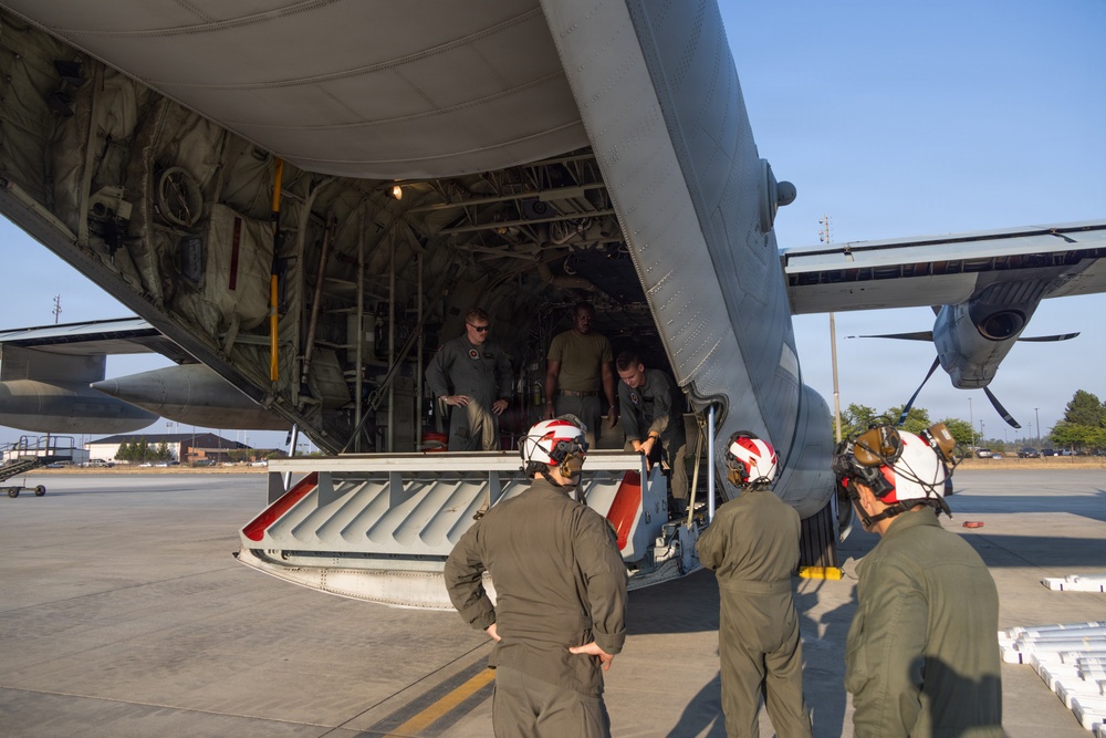 Military personnel are gathered at the open rear cargo ramp of a large military transport aircraft, likely a C-130 Hercules, on a flight line or tarmac. Several crew members wearing flight suits and white helmets with hearing protection are standing outside examining or loading equipment, while others stand inside the aircraft's cargo bay. A piece of specialized equipment with red and silver components is positioned at the base of the ramp.