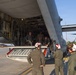 Military personnel are gathered at the open rear cargo ramp of a large military transport aircraft, likely a C-130 Hercules, on a flight line or tarmac. Several crew members wearing flight suits and white helmets with hearing protection are standing outside examining or loading equipment, while others stand inside the aircraft's cargo bay. A piece of specialized equipment with red and silver components is positioned at the base of the ramp.