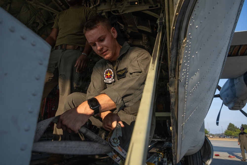 A young military service member in an olive/sage green flight suit is working inside the open door or hatch of a military aircraft. He is focused on manipulating equipment or hardware components within the aircraft's interior. A second person in similar uniform is partially visible in the background.