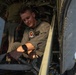 A young military service member in an olive/sage green flight suit is working inside the open door or hatch of a military aircraft. He is focused on manipulating equipment or hardware components within the aircraft's interior. A second person in similar uniform is partially visible in the background.