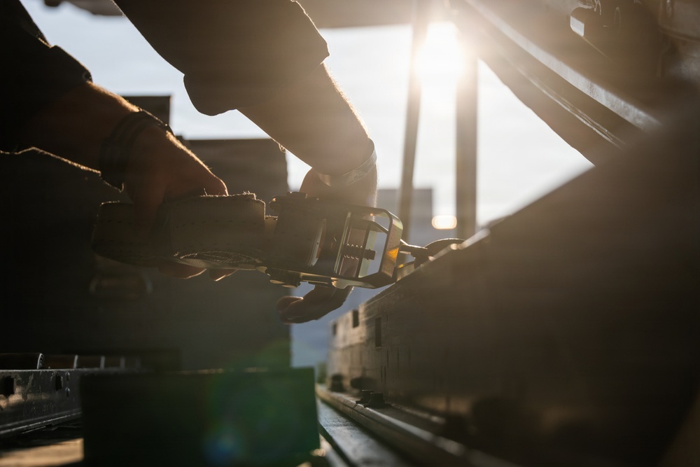 A person wearing work gloves is operating a ratchet strap or tie-down mechanism, likely securing cargo on a flatbed truck or trailer. The image is captured at low angle during golden hour, with strong backlighting from the sun creating dramatic lens flare and silhouette effects. Industrial or transportation infrastructure is visible in the background.