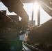 A person wearing work gloves is operating a ratchet strap or tie-down mechanism, likely securing cargo on a flatbed truck or trailer. The image is captured at low angle during golden hour, with strong backlighting from the sun creating dramatic lens flare and silhouette effects. Industrial or transportation infrastructure is visible in the background.