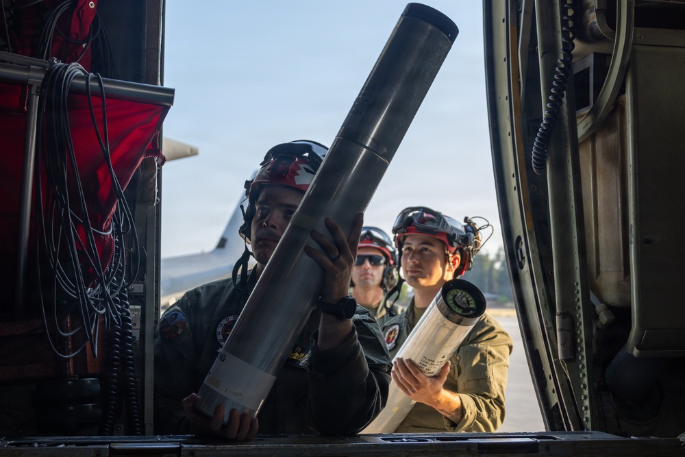 Military personnel wearing flight suits and helmets with red cross/maple leaf insignia are loading cylindrical tube-shaped objects into what appears to be a military helicopter or aircraft. Three crew members are visible, with two actively handling the cylindrical containers while a third observes. The setting appears to be a tarmac or airfield, with aircraft visible in the background.