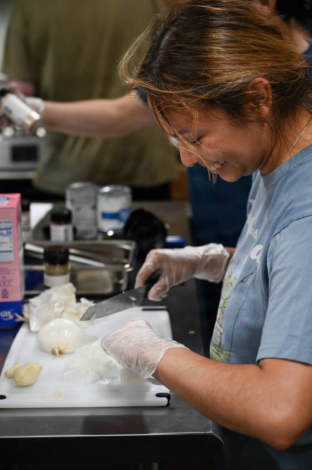 A woman wearing disposable gloves and a blue t-shirt is smiling while chopping onions on a white cutting board in what appears to be a commercial or community kitchen. She is using a large chef's knife and has several onions on the board, some already diced. In the background, another person is also working at the same counter surrounded by various kitchen supplies and condiments.