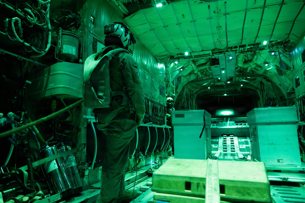 The image shows the interior of a military cargo aircraft illuminated by green lighting, consistent with night vision operations or low-light tactical conditions. One or more personnel in flight suits and helmets are visible standing among cargo containers and equipment secured to the aircraft floor. The scene depicts an active military airlift or logistics operation.
