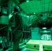 The image shows the interior of a military cargo aircraft illuminated by green lighting, consistent with night vision operations or low-light tactical conditions. One or more personnel in flight suits and helmets are visible standing among cargo containers and equipment secured to the aircraft floor. The scene depicts an active military airlift or logistics operation.