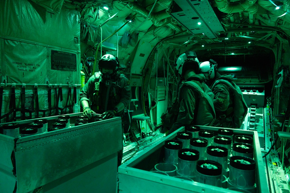 The interior of a military aircraft illuminated by green lighting, consistent with night operations or NVG (night vision goggle) compatible red/green lighting protocols. Two or more military personnel wearing flight helmets and oxygen masks are working with rows of cylindrical canisters arranged in metal trays or racks. The scene appears to depict an airborne mission involving the preparation or management of dispensable payloads, likely sonobuoys or similar air-dropped devices.