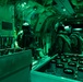 The interior of a military aircraft illuminated by green lighting, consistent with night operations or NVG (night vision goggle) compatible red/green lighting protocols. Two or more military personnel wearing flight helmets and oxygen masks are working with rows of cylindrical canisters arranged in metal trays or racks. The scene appears to depict an airborne mission involving the preparation or management of dispensable payloads, likely sonobuoys or similar air-dropped devices.