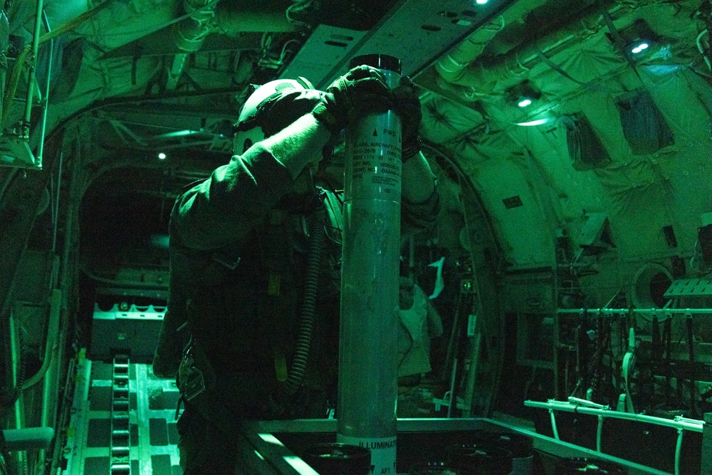 A military aircrew member is working inside what appears to be a large military transport or gunship aircraft, illuminated by green lighting. The individual is handling a cylindrical munition or flare canister, with visible labeling including the word 'ILLUMINAT[ION]' at the base. The green ambient lighting suggests night operations or use of night-vision-compatible lighting systems.