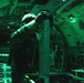 A military aircrew member is working inside what appears to be a large military transport or gunship aircraft, illuminated by green lighting. The individual is handling a cylindrical munition or flare canister, with visible labeling including the word 'ILLUMINAT[ION]' at the base. The green ambient lighting suggests night operations or use of night-vision-compatible lighting systems.