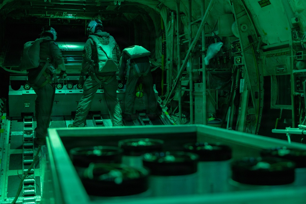 The image shows the interior of a military cargo aircraft, likely a C-130 or similar transport, illuminated by green lighting. Two personnel in flight suits and helmets are visible inside the cargo bay, appearing to conduct loading or inspection activities. The cargo floor features roller conveyors and tie-down tracks typical of military transport aircraft.