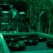 The image shows the interior of a military cargo aircraft, likely a C-130 or similar transport, illuminated by green lighting. Two personnel in flight suits and helmets are visible inside the cargo bay, appearing to conduct loading or inspection activities. The cargo floor features roller conveyors and tie-down tracks typical of military transport aircraft.