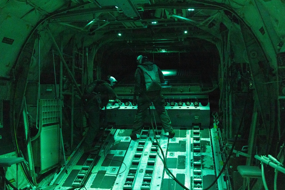 The image shows the interior of a military cargo aircraft, likely a C-130 Hercules, illuminated by green night-vision-compatible lighting. Two personnel wearing flight helmets and tactical gear are visible on the cargo floor, appearing to conduct pre-drop or loading operations. The cargo bay is empty of freight, with the roller-equipped floor track system clearly visible.