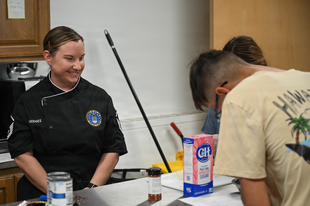 A smiling woman wearing a black chef's jacket with a U.S. Air Force emblem and the name 'HODGES' is standing at a kitchen counter. She appears to be instructing or supervising individuals in a cooking class or culinary training setting. A box of C&amp;H Pure Cane Sugar and other cooking ingredients are visible on the counter.