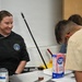 A smiling woman wearing a black chef's jacket with a U.S. Air Force emblem and the name 'HODGES' is standing at a kitchen counter. She appears to be instructing or supervising individuals in a cooking class or culinary training setting. A box of C&amp;H Pure Cane Sugar and other cooking ingredients are visible on the counter.