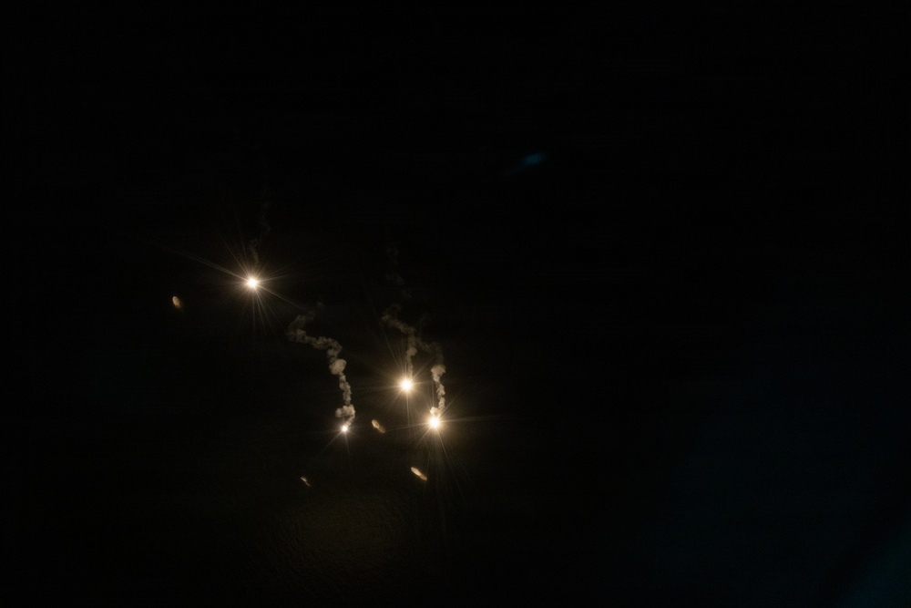 A nighttime photograph showing multiple bright light sources against a nearly completely dark background. Several luminous points are visible with smoke or vapor trails rising from some of them, suggesting pyrotechnic or flare activity. The image is predominantly dark with the light sources concentrated in the left-center portion of the frame.